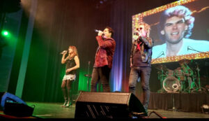 Ally Fowler, Scott Carne and Brian Mannix standing beside each other on stage singing a song together.
