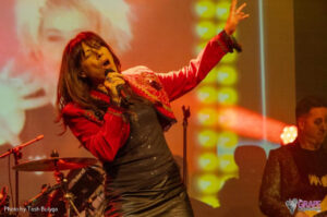Ally Fowler wearing a red bolero over a black top and black leather skirt as she sings into a microphone.
