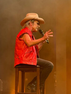 Brian Mannix sitting on a chair pretending to be Molly Meldrum while weating a red vest and Akubra hat.