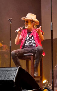 Brian Mannix sitting on a chair pretending to be Molly Meldrum while weating a red vest and Akubra hat.