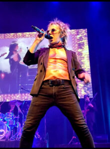 Brian Mannix singing on stage with an open jacket and a T-shirt with muscly abs printed on it.