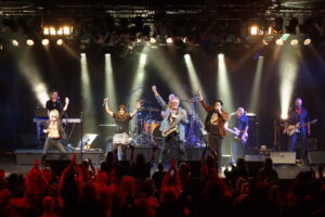 Brian Mannix, Ally Fowler, Wilbur Wilde and Scott Carne with their arms in the air at the end of a song that they are singing together. The band is in the background featuring a keyboard player, drummer, bass guitarist and guitarist.The crowd in front of them is cheering.