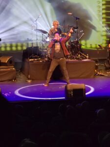 Scott Carne on stage surrounded by blue glowing lights. He's wearing a Little Richard shirt, red jacket and black jeans. He's holding a microphone with one arm in the air while singing.