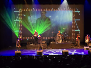 Scott Carne and Ally Fowler singing together on stage with a band behind them and green neon lighting.