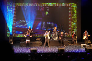 Scott Carne, Ally Fowler and Brian Mannix singing on stage with a band behind them and blue lights shining up.
