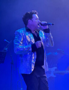 Scott Carne wearing a glittery gold jacket holding a microphone singing on stage.