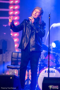 Sean Kelly holding a microphone singing on stage with blue lighting.