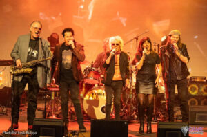 Wilbur Wilde, Scott Carne, Brian Mannix and Ally Fowler singing on stage with orange lighting.